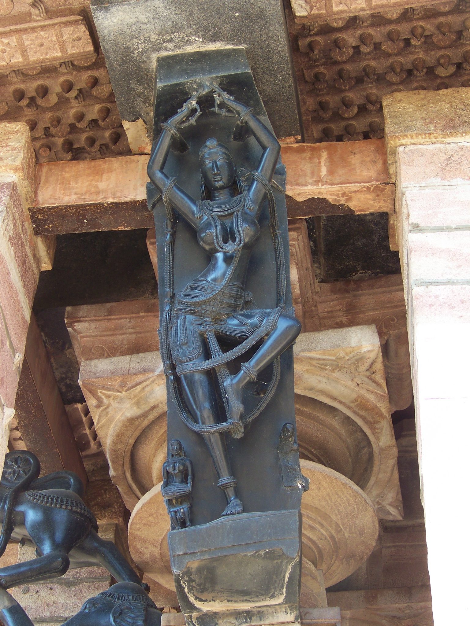 A single dark stone bracket sculpture of a woman with arms raised overhead, set on a column capital beneath the eave of the Ramappa mandapa