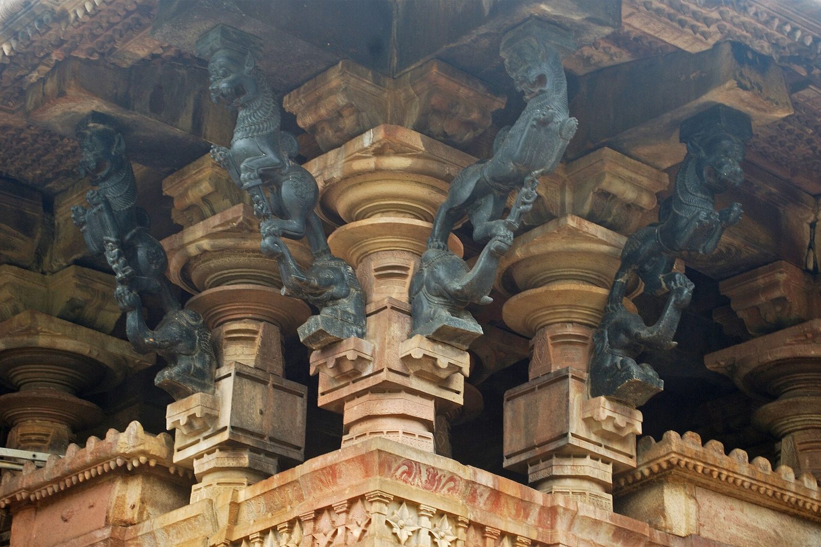 Dark stone bracket sculptures of rearing lion-like beasts standing over small elephants, set on the capitals of sandstone columns beneath the eave of the Ramappa mandapa