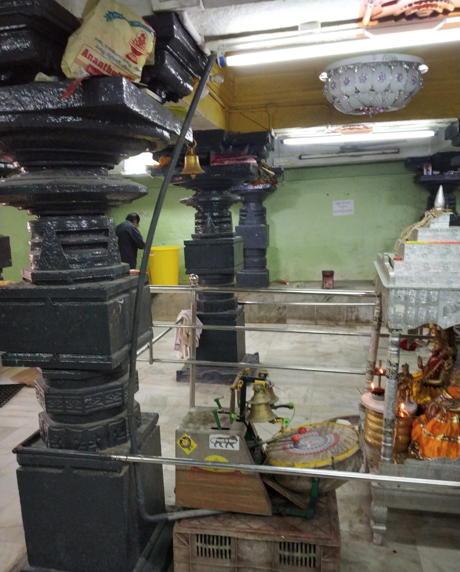 Inside a temple hall, heavy dark stone pillars stand on square bases. Their shafts are carved in stacked, rounded and square layers. The room has a tiled floor, a painted green wall, tube lights and a chandelier. Festival images dressed in orange stand on a silver-covered stand at the right.
