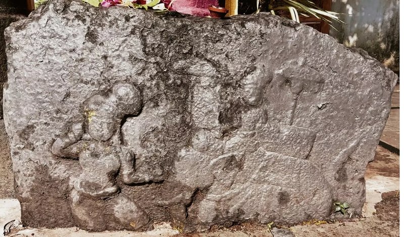 A weathered slab of dark stone with worn carvings in low relief. The carving on the left appears to show a kneeling person, and other worn shapes fill the right side. Flowers lie on top of the slab.