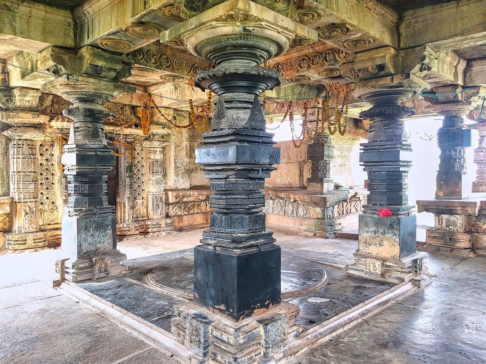 Inside a temple hall, a central pillar of dark polished stone with many rounded and square sections stands on a square base. More dark pillars and carved granite beams surround it.