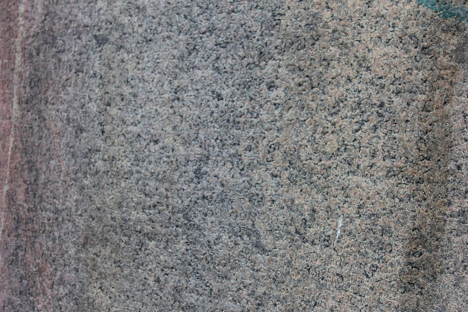 A close view of speckled granite with rows of small, shallow, rounded letters cut into it. A reddish band runs down the left edge.