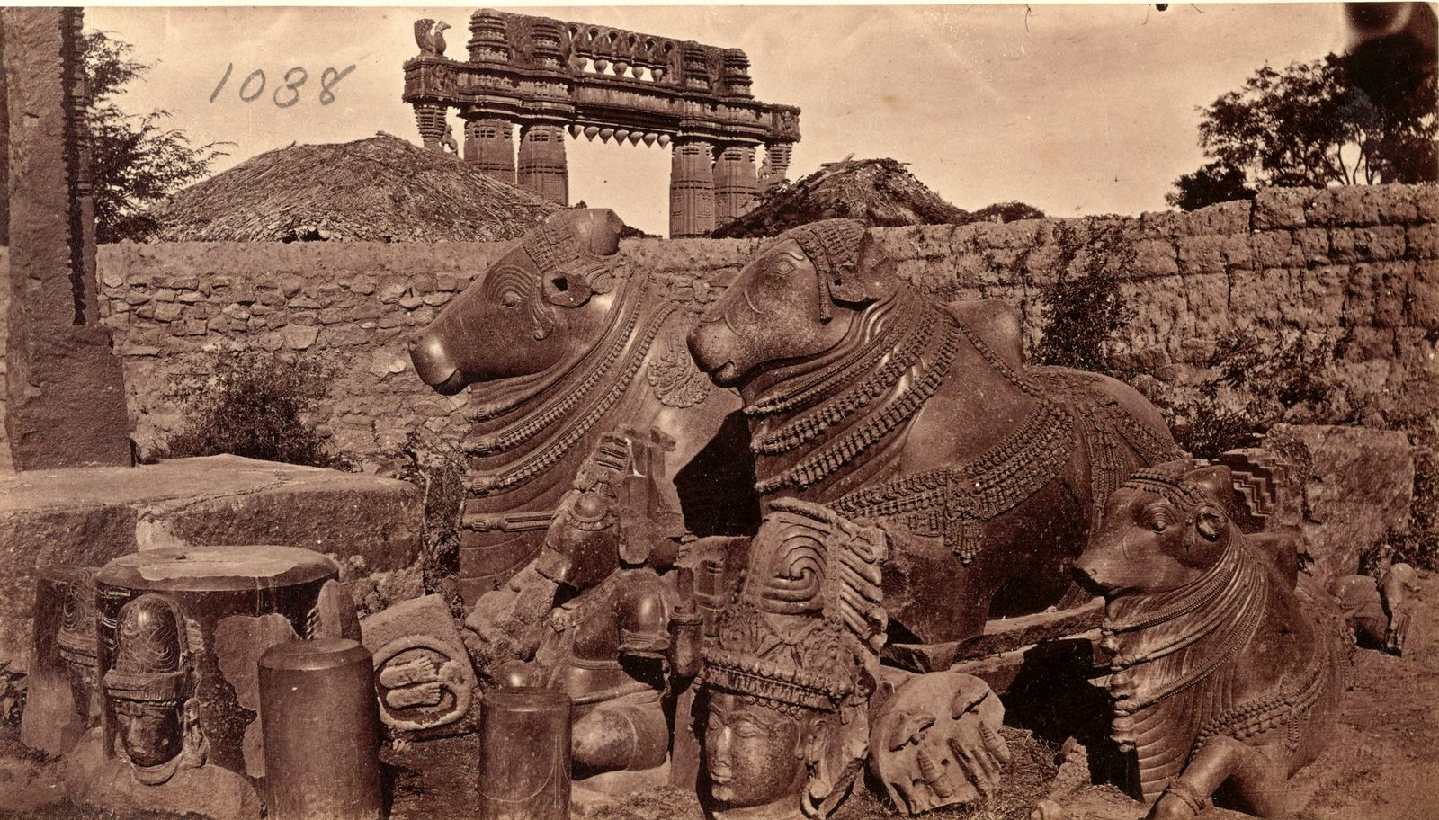 Two large carved stone bulls lie side by side among broken carved stones. A rough stone wall stands behind them. The top of a carved stone gateway rises above the wall.
