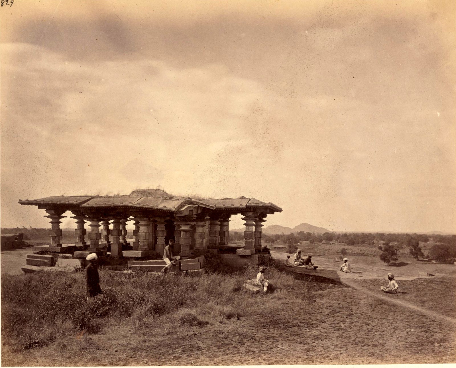 A flat-roofed stone pavilion on many pillars stands on a slight rise in open grassland. Several people sit and stand around it. Hills show on the horizon.