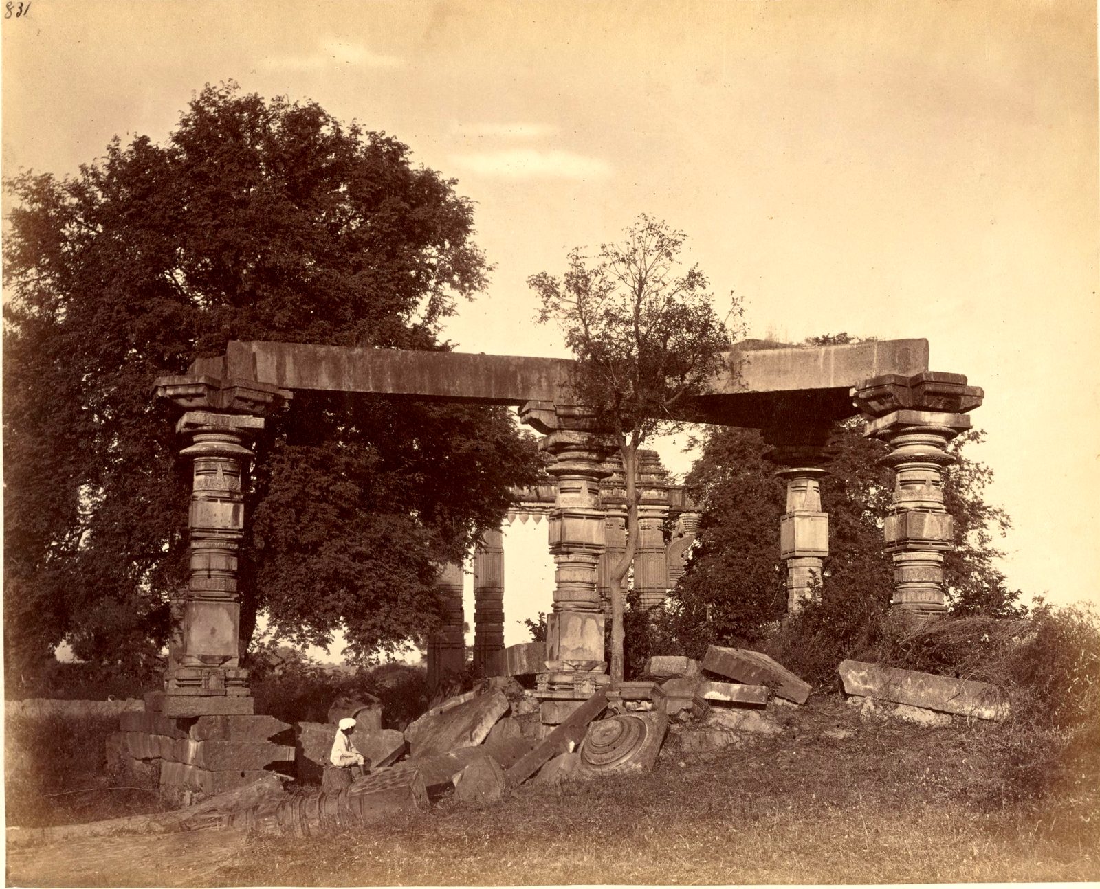 Four carved stone pillars hold up a broken stone beam among large trees. Fallen carved blocks lie on the ground. A man sits among the fallen stones.