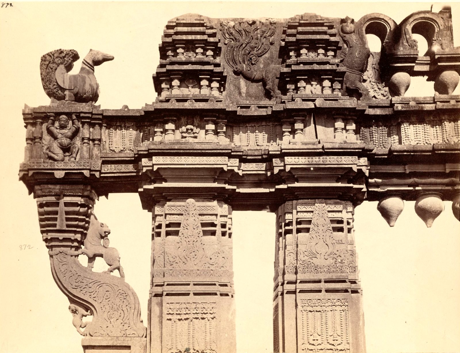 Close view of the top of a carved stone gateway. A stone swan sits on the end of the crossbeam, and a carved bracket with a rearing animal supports it. Hanging stone buds line the underside of the beam.