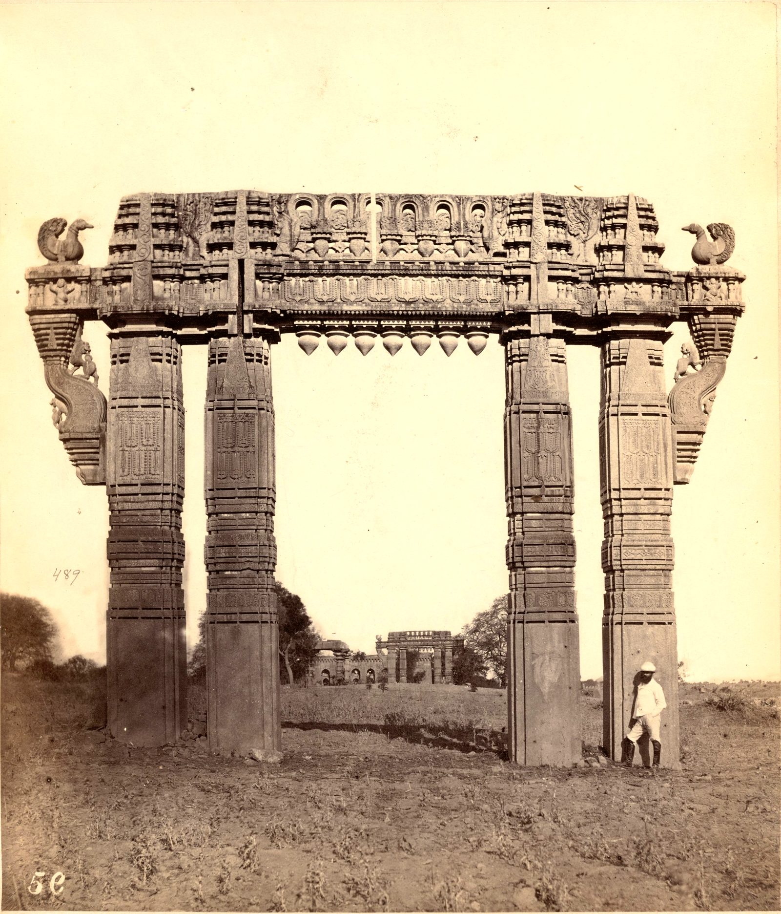A tall stone gateway of four carved pillars and a carved crossbeam stands in open ground. Stone swans sit on the ends of the crossbeam. A long building with arches shows through the gateway, and a man stands at the right pillar.