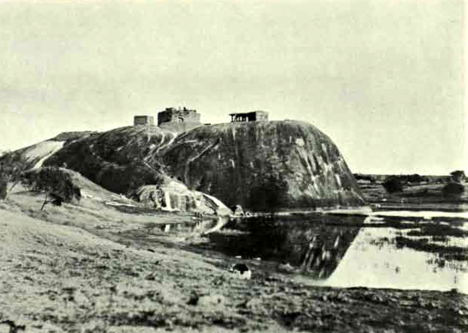 A large bare granite rock rises beside a pool of water. Small buildings stand on its top. Grass covers the slope in front.