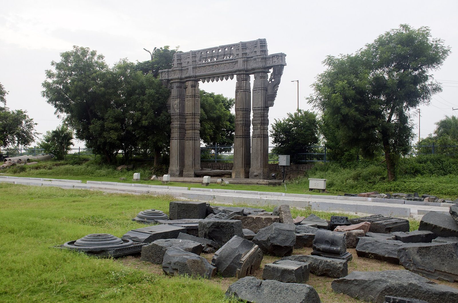 A keerthi thorana gateway at Warangal standing in parkland, with fallen carved stone blocks and column bases piled on the grass in front