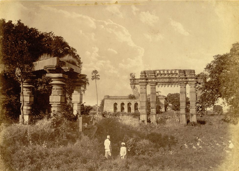 Sepia photograph of overgrown ruins: a pillared structure at left, a stone gateway at right and an arcaded building in the distance, with two people standing in the scrub